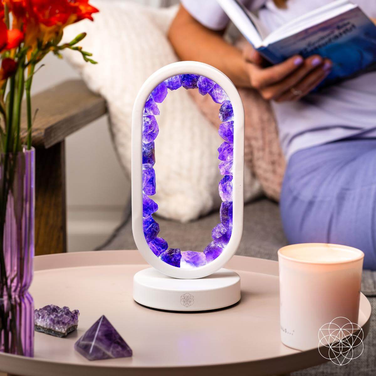 Decorative lamp with purple crystals on a table next to a vase of flowers and a candle, with a person reading a book in the background.